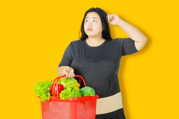 Elhízott Töpreng Miközben Egy Bevásárlókocsit Tart Zöldséggel Stúdióban Sárga Háttérrel — Stock Fotó