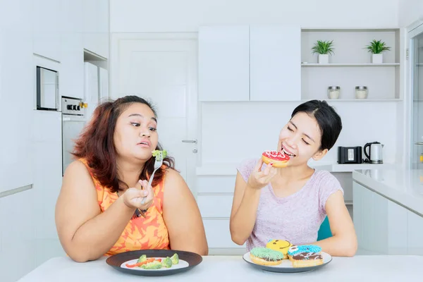 Lose weight concept. Unhappy fat woman eating salad while mocked by her friend with eating donuts in the kitchen at home