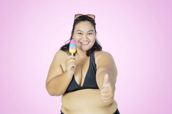 カメラに親指を立てながらアイスクリームを持ちピンクの背景を持つスタジオに立つ太った女 — ストック写真