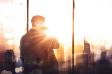 Erkek müdürün bir fincan sıcak kahvenin keyfini çıkarırken, gün batımında arka planda pencerenin önünde dikilirken...