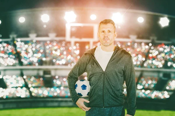 Soccer coach man holding a ball while standing in the stadium with spectators seat background. Shot at night time