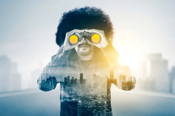 Double exposure of young Afro man looking through a binocular while standing with cityscape background