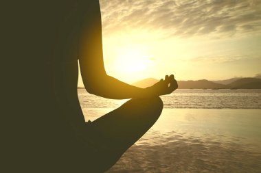 Gün batımında arka planda kıyı şeridinde otururken yoga yapan genç bir kadının silueti.