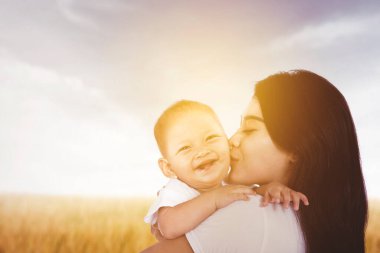Mutlu anne bebeğini kucağına alıp öperken arka planda güneş ışığıyla çayırda dikiliyor. Sonbahar zamanı çekilmiş.