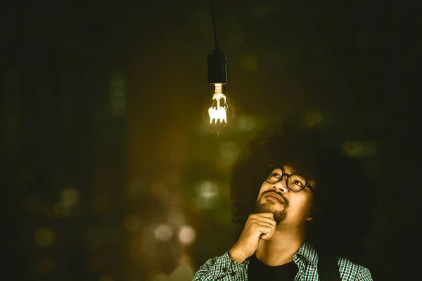 Male college student thinking an idea while looking at a bright bulb over his head and standing with blurred sparkling lights background
