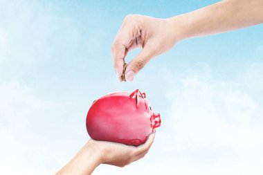 Bilinmeyen bir el, mavi gökyüzü arkaplanlı bir kumbara içine para sokuyor.