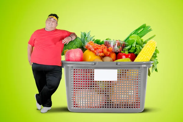胖男人靠在满是蔬菜的购物车上 站在有绿色背景的工作室里 — 图库照片