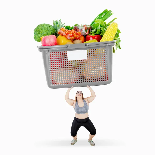 スタジオで野菜や果物でいっぱいの箱を持ち上げながらスポーツウェアを着た太った女性 白地に隔離された — ストック写真