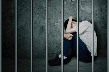 Close up of young man looks sad while sitting behind bars in the jail