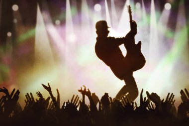 Elektro gitar çalan erkek gitaristin silueti, hayranlarının elinin yakınındaki konser sahnesinde duruyor.