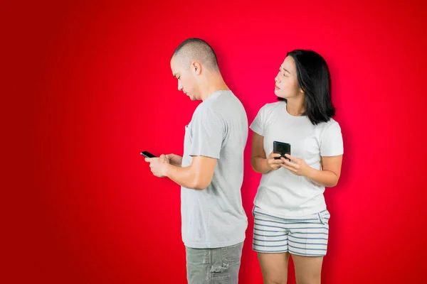 Asian young woman spying at cellphone of her husband while standing together in the studio with red background