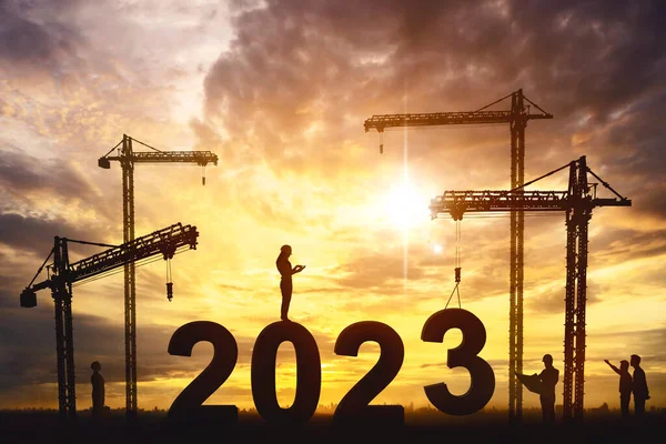 Group silhouette of engineers assembling 2023 number with cranes while working together in construction site with dusk sunlight background