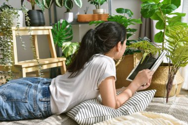 Oturma odasında dinlenen ve okuyan bir kadın, yapay bitki, keman yaprağı incir ağacı, ev içi ve hava temizliği için tropikal doğal ev bitkisi.