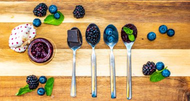 Blueberry, blackberry and chocolate are placed in silver spoon on wooden desk. Ingredients for marmalade. Fresh fruit around.