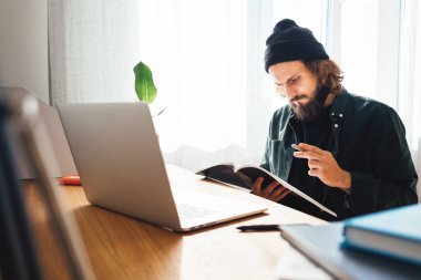 Online eğitim. Dizüstü bilgisayarla çevrimiçi konferans dinlerken üniversite öğrencisi çalışıyor. Sakallı hippi ev işi yapıyor.