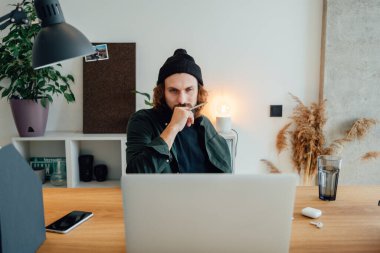 Online eğitim. Dizüstü bilgisayarla çevrimiçi konferans dinlerken üniversite öğrencisi çalışıyor. Sakallı hippi ev işi yapıyor.