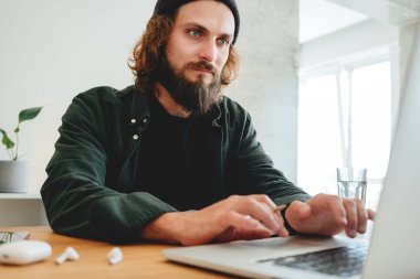 Online eğitim. Dizüstü bilgisayarla çevrimiçi konferans dinlerken üniversite öğrencisi çalışıyor. Sakallı hippi ev işi yapıyor.