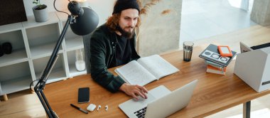 Online eğitim. Dizüstü bilgisayarla çevrimiçi konferans dinlerken üniversite öğrencisi çalışıyor. Sakallı hippi ev işi yapıyor. Geniş resim