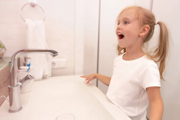 Klein gelukkig meisje, Europees uiterlijk, wast in de badkamer in de ochtend. Kijk in de spiegel en lacht.. — Stockfoto