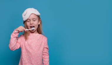 Pijamalı, gözleri kapalı küçük bir kız mavi arka planda dişlerini fırçalıyor.