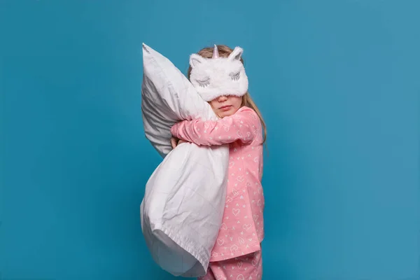 Girl in pajamas and with an eye mask stands on a blue background with a pillow