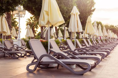 Şafakta havuz kenarında boş plastik güneşlikler ve kapalı şemsiyeler.