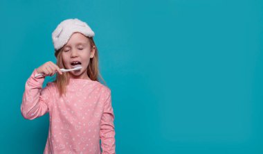 Pijamalı, gözleri kapalı küçük bir kız mavi arka planda dişlerini fırçalıyor.