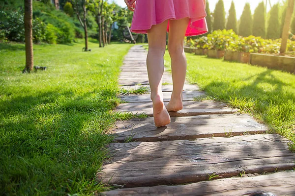 A small, thin girl in a pink dress runs along the path on a sunny day. Close-up of small feet. The concept of childrens feet health, prevention of flat feet, hallux valgus, massage, wellness.