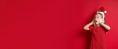 Uzun afiş, kırmızı arkaplan ve kopyalama alanı. Mutlu Noeller ve mutlu yıllar. Noel Baba şapkalı, kulaklıklı, mutlu bir şekilde gülen küçük sarışın, beyaz bir kadın.