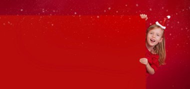 Uzun afiş, kırmızı arkaplan ve kopyalama alanı. Mutlu Noeller ve mutlu yıllar. Noel Baba şapkalı küçük sarışın, beyaz kadın, mutlu bir şekilde gülüyor, büyük bir kağıt parçasının arkasından dışarı bakıyor.