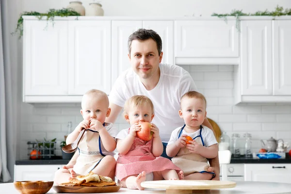 A man holds and hugs three babies in his arms. Happy father of triplets in front of a white kitchen. The concept of a happy family, eco, healthy food. Father on parental leave