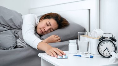 Asyalı kadın kendini hasta hissediyor ve vücut ısısını ölçmek ve ilaç almak için termometre kullandıktan sonra yatakta battaniyede yatarken masadaki ilaçlara dokunmak için ellerini kullanıyor.