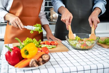 Asyalı romantik çift ahşap kesim tahtasına maşa ve domates koyarken genç adam kepçeyle evde sağlıklı yemek pişirmek için sebze salatası hazırlıyor.