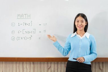 Beyaz tahtanın önünde duran ve e-öğrenme sınıfında canlı yayın videosuyla konuşurken yeni dersi açıklamaya yarayan matematik denklemleri yazan kadın öğretmen.