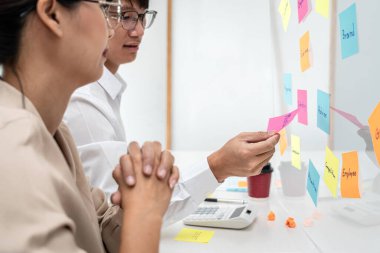 İş kadını yeni bir başlangıç projesi hakkında düşünceli ve ortakla pazarlama planı planlama konusunda beyin fırtınası yaparken iş adamı ofisteki cam bir duvarda fikrini paylaşmak için notlar tutuyor.