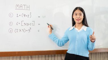 Beyaz tahtanın önünde duran ve e-öğrenme sınıfında canlı yayın videosuyla konuşurken yeni dersi açıklamaya yarayan matematik denklemleri yazan kadın öğretmen.