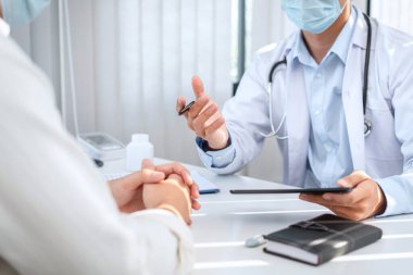 Tıbbi maske takan erkek doktor, klinikteki hastaya muayene sonuçları hakkında rapor verdikten sonra belirtileri, tedaviyi ve tavsiyeyi açıklamak için tablet tutuyor.
