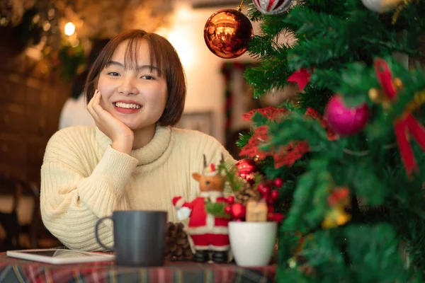 セーターを着た美しい女性は笑顔で 輝くボールで飾られたクリスマスツリーの近くのコーヒーカップと椅子に座ってクリスマスやリビングルームで新年を祝うための装飾 — ストック写真