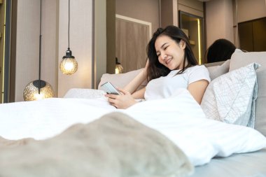 Sosyal medyada sohbet etmek ve film izlemek için akıllı telefon kullanan Asyalı bir kadın. Yatak odasındaki yatağa uzanıp hafta sonunun tadını çıkarıyor.