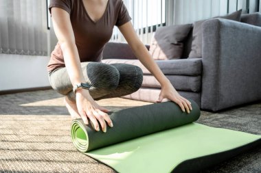 Spor kıyafetli genç bir kadın paspası kaldırıyor ve evdeki oturma odasında yoga egzersiz dersi için dizüstü bilgisayar hazırlıyor..