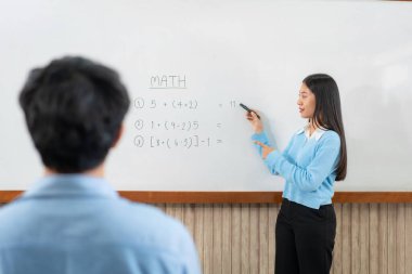 Kadın öğretmen beyaz tahtanın önünde duruyor ve matematik dersinde öğrencilere açıklama yapmak için güverteye matematik denklemleri yazıyor.