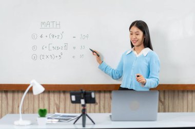 Kadın öğretmen beyaz tahtanın önünde duruyor ve yeni dersi açıklamaya matematik denklemleri yazıyor. Kamera ise canlı yayın videosuyla e-öğrenim sınıfında konuşuyor.