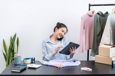 Kadın moda tasarımcısı tabletteki siparişleri kontrol ediyor ve evdeki müşteriye teslimat için karton kutularda kıyafet hazırlıyor..