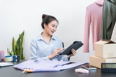 Kadın moda tasarımcısı tabletteki siparişleri kontrol ediyor ve evdeki müşteriye teslimat için karton kutularda kıyafet hazırlıyor.