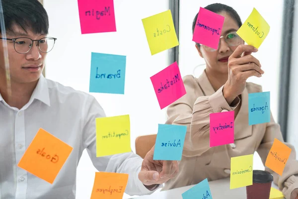 ガラスの壁にキーワードを押した2人のビジネス人がオフィスで一緒にマーケティング計画を計画するための新しいビジネスプロジェクトについてブレインストーミングする — ストック写真