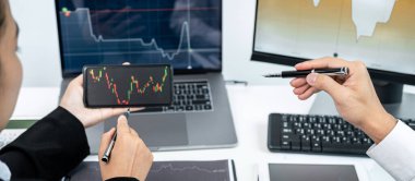 İki iş arkadaşı akıllı telefondaki veri tablosunu veri borsasını kontrol etmek ve birlikte dizüstü bilgisayardan bilgi ararken piyasadaki oyun hisselerini tartışmak için işaret ediyor.