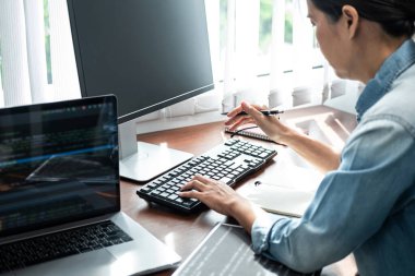Asyalı kadın programcı elinde kalem tutarken ve dizüstü bilgisayardaki yeni proje hakkında kod verileri yazmak için klavyeye bakarken aynı zamanda web sitesi veya yazılım geliştirme ofisindeki uygulamalar için kod yazmaya çalışıyor