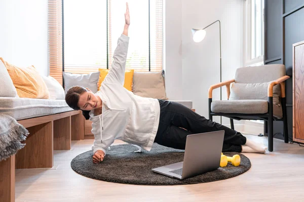 Asyalı kadın yoga egzersizi yapıyor, kolunu kaldırıyor ve dizüstü bilgisayarla evdeki oturma odasında yoga dersi veriyor.