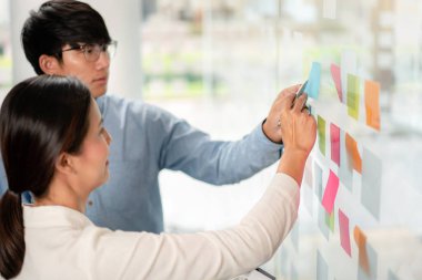 İki işletme yöneticisi, ofiste birlikte pazarlama planının planlanmasına yönelik yeni bir iş projesi hakkında fikir alışverişinde bulunmak için kilit noktayı duvara yazıp not alıyorlar..