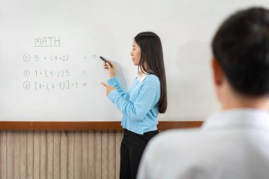 Kadın öğretmen beyaz tahtanın önünde duruyor ve matematik dersinde öğrencilere açıklama yapmak için güverteye matematik denklemleri yazıyor.